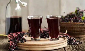 Dwa kieliszki ciemnego, aromatycznego soku z czarnego bzu stoją obok butelki i świeżych owoców.