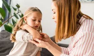 Mama opatruje łokieć córki, na którym widać zaczerwienienie i otarcie. Dziecko ma zamknięte oczy, jakby bolało je to **trudno gojące się rany**.