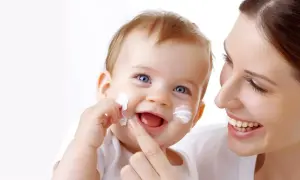 Uśmiechnięta mama aplikuje Sudocrem na policzki dziecka, chroniąc je przed podrażnieniami i grzybicą.
