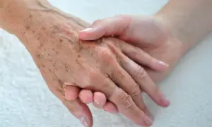 Dwie dłonie, starsza z widocznymi plamami starczymi i młodsza, trzymają się nawzajem.
