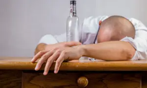 Mężczyzna w białej koszuli leży na stole, głowa schowana w ramionach. Obok pusta butelka, symbolizująca skutki nadużycia alkoholu, jakimi mogą być wymioty po alkoholu.