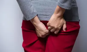 Osoba w czerwonych spodniach i szarej bluzce drapie się w okolicy odbytu. Szuka ulgi, zastanawiając się, czym smarować odbyt przy owsikach.