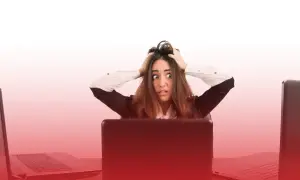 Kobieta z przerażeniem w oczach, rękami wplątanymi we włosy, siedzi przy biurku otoczona laptopami. To obraz stresu w pracy.