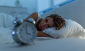 Zmartwiona kobieta leży w łóżku, patrząc na budzik. To znak, że czas pomyśleć, jak walczyć z bezsennością.