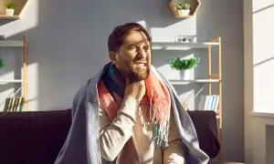 Mężczyzna z zarostem, okryty kocem i szalikiem, odczuwa ostry ból gardła przy przełykaniu, choć nie ma gorączki.