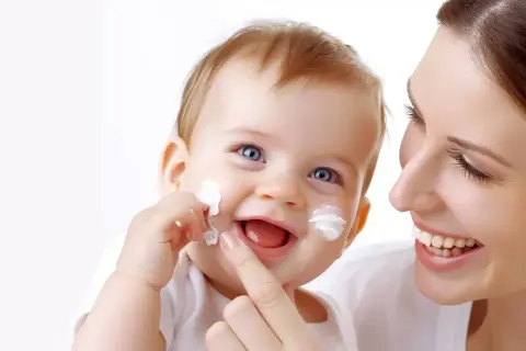 Uśmiechnięta mama aplikuje Sudocrem na policzki dziecka, chroniąc je przed podrażnieniami i grzybicą.