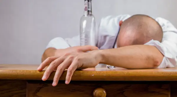 Mężczyzna w białej koszuli leży na stole, głowa schowana w ramionach. Obok pusta butelka, symbolizująca skutki nadużycia alkoholu, jakimi mogą być wymioty po alkoholu.