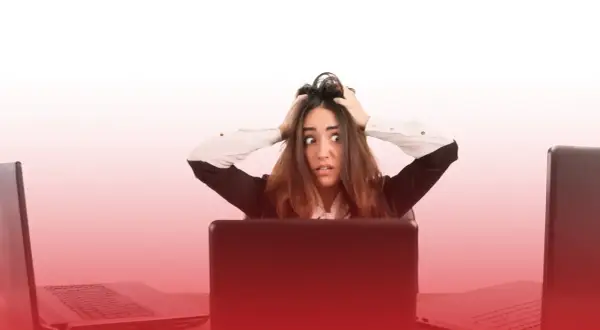 Kobieta z przerażeniem w oczach, rękami wplątanymi we włosy, siedzi przy biurku otoczona laptopami. To obraz stresu w pracy.