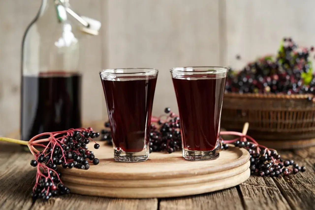 Dwa kieliszki ciemnego, aromatycznego soku z czarnego bzu stoją obok butelki i świeżych owoców.