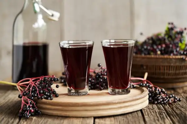 Dwa kieliszki ciemnego, aromatycznego soku z czarnego bzu stoją obok butelki i świeżych owoców.