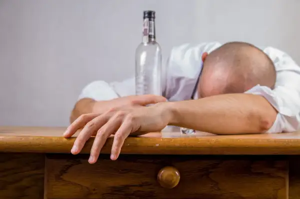 Mężczyzna w białej koszuli leży na stole, głowa schowana w ramionach. Obok pusta butelka, symbolizująca skutki nadużycia alkoholu, jakimi mogą być wymioty po alkoholu.