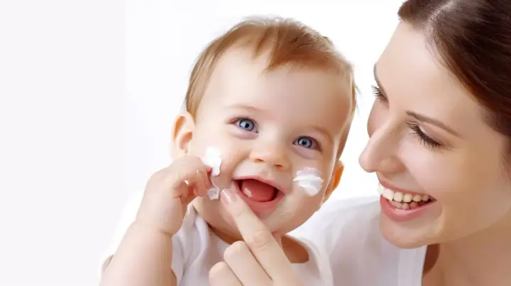 Uśmiechnięta mama aplikuje Sudocrem na policzki dziecka, chroniąc je przed podrażnieniami i grzybicą.